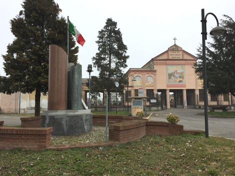 Monumento ai caduti di Belgioioso con bandiera italiana e chiesa sullo sfondo.