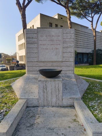 Monumento ai caduti militari con un design sobrio, decorato da un vaso nero e circondato da verde.