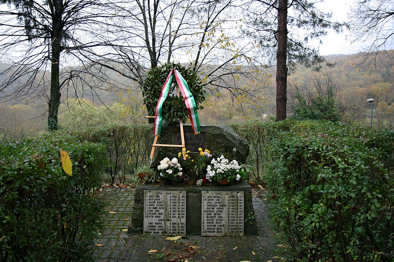 Monumento ai caduti di Polcanto, circondato da fiori e piante, in un contesto naturale sereno.