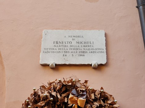 Lapide in pietra con iscrizione, fissata su una parete intonacata sopra una corona di fiori secchi.