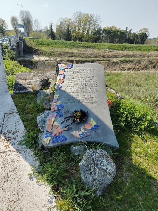 Lastra commemorativa dedicata agli sminatori, adornata con fiori e colori, immersa in un paesaggio naturale.