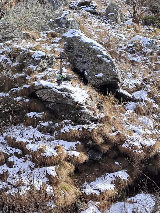 Piccola croce in metallo tra le rocce di un pendio erboso innevato.