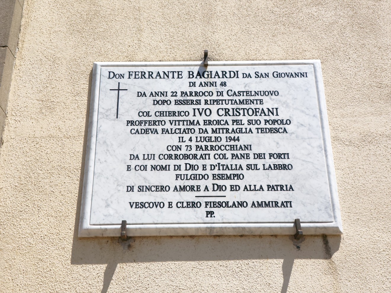 Lapide in marmo con iscrizione e croce incisa, fissata su una parete esterna intonacata.