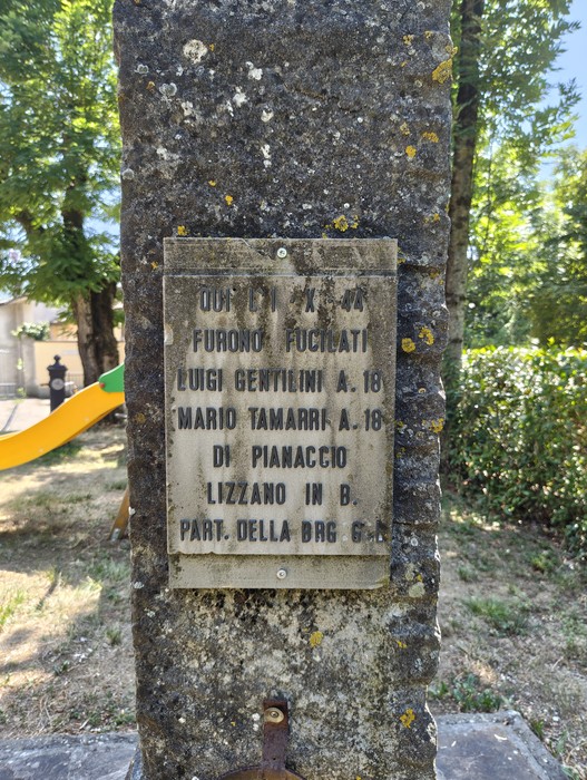 Targa in pietra con iscrizione, fissata su un pilastro in cemento all'aperto, tra alberi e vegetazione.