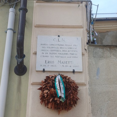 Lapide in marmo con iscrizione, fissata su parete esterna, con corona di foglie secche appesa sotto.
