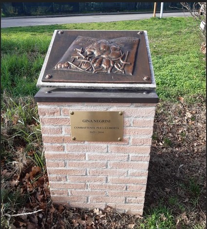 Cippo commemorativo dedicato a Gina Negrini, con rilievo artistico e placca informativa, circondato da verde.