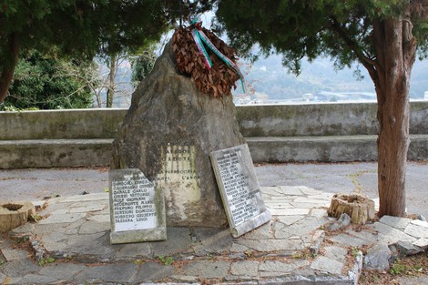 Monumento ai caduti di Morego, con una roccia centrale e lastre commemorative sotto alberi verdi.