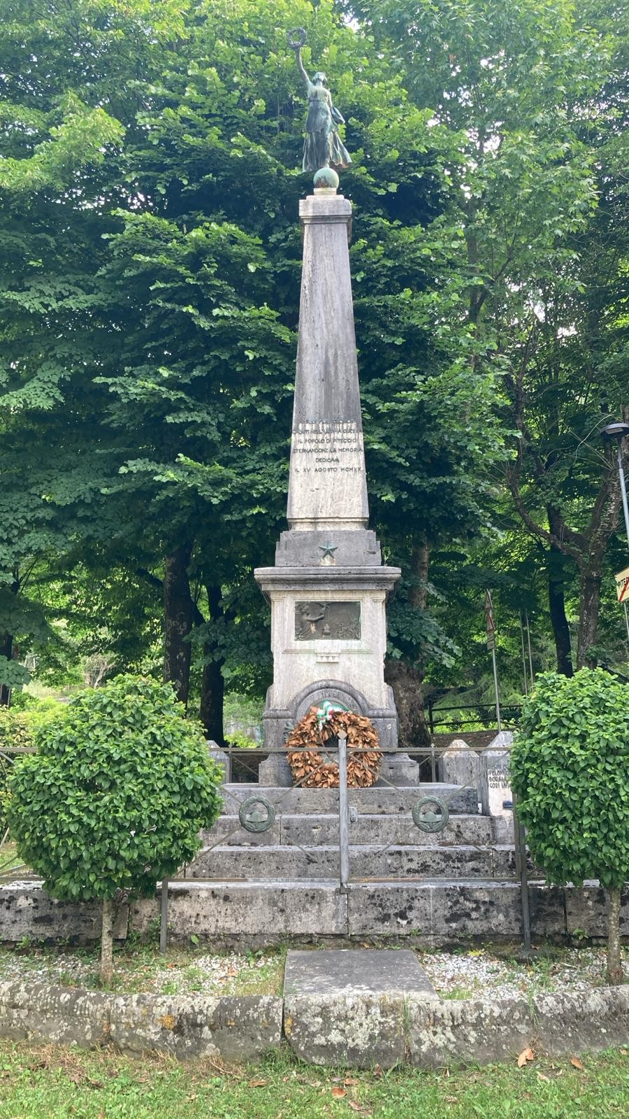 Monumento ai caduti di Piteccio, con obelisco e decorazioni floreali, immerso nel verde.