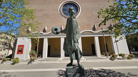 Monumento a Minzoni, statua in bronzo con figura stilizzata, sullo sfondo un edificio con grande finestra circolare.