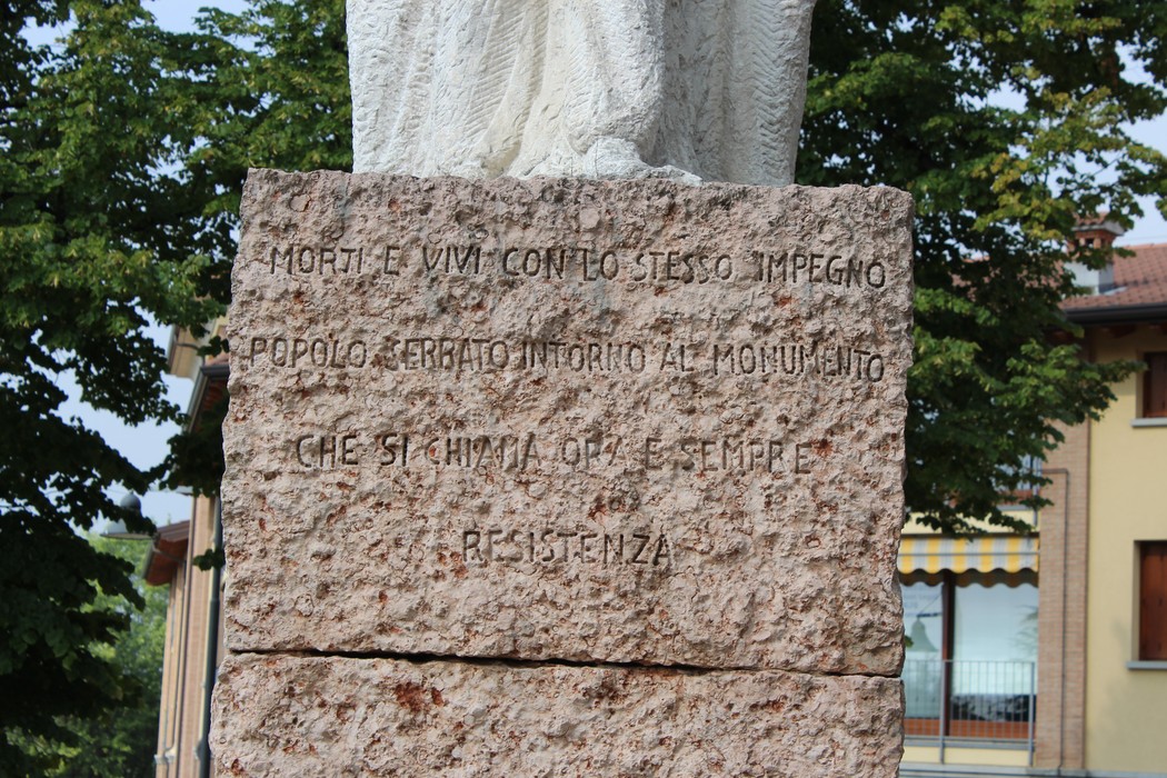 Monumento alla Resistenza, con base in pietra e statua, circondato da alberi e edifici.