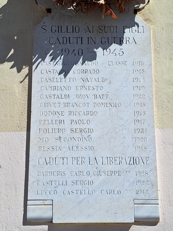 Lapide in marmo con lungo elenco di nomi, fissata su una parete esterna, sotto un ramo di piante.