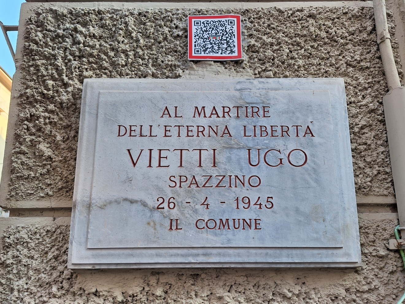 Lapide in marmo con iscrizione su parete esterna intonacata, con QR code applicato sopra.