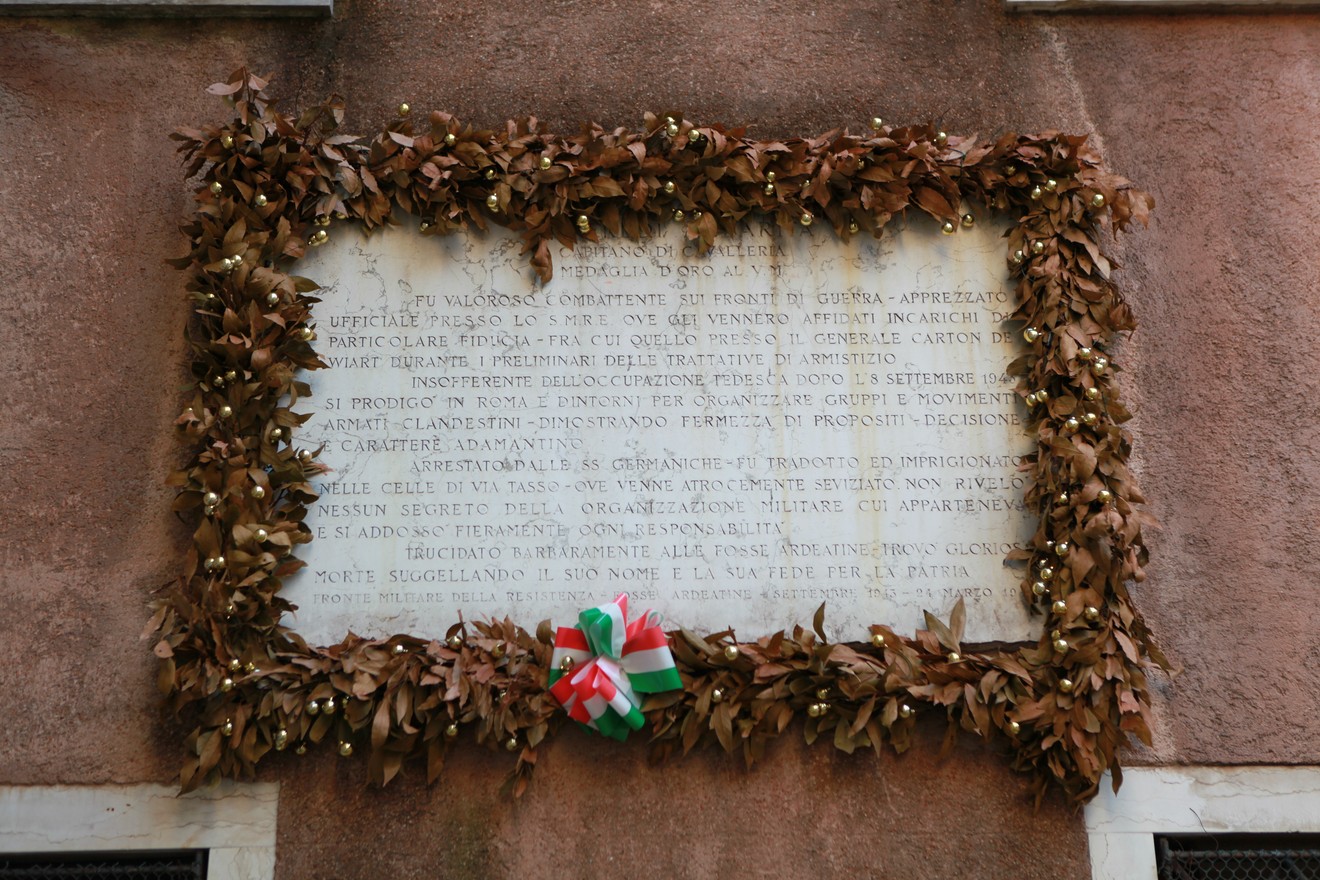 Lapide commemorativa adornata con foglie e un nastro tricolore su una parete.