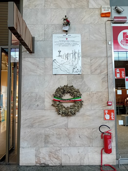 Lapide commemorativa ai deportati con corona di alloro e decorazioni, collocata su una parete di pietra.