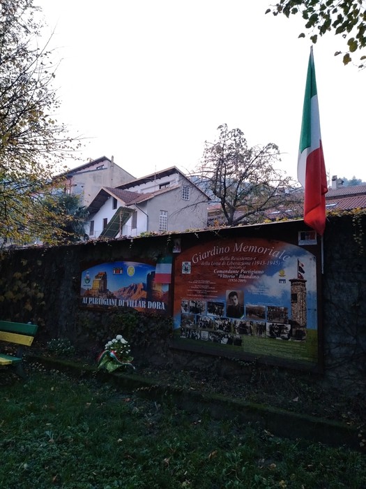 Giardino memoriale a Blandino, con manifesti commemorativi e la bandiera italiana in un contesto naturale e tranquillo.