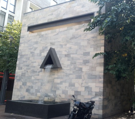 Monumento a Pertini in pietra, con fontana triangolare e iscrizione, addossato a una parete in una piazza.