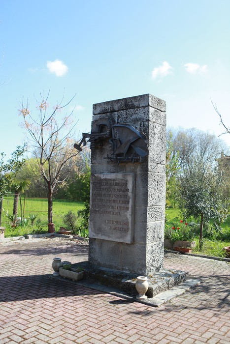 Monumento ai caduti del Tiro a segno nazionale, con iscrizione su pietra e un albero dietro. Atmosfera serena e naturale.