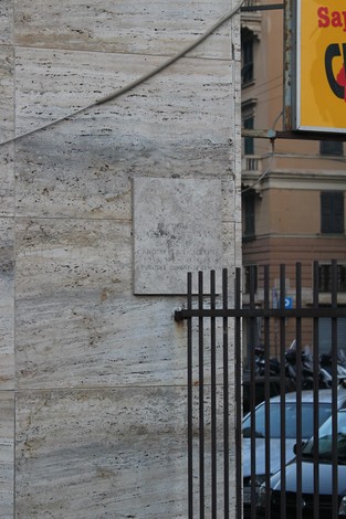 Lapide commemorativa su un muro di pietra, in ambiente urbano con veicoli parcheggiati.