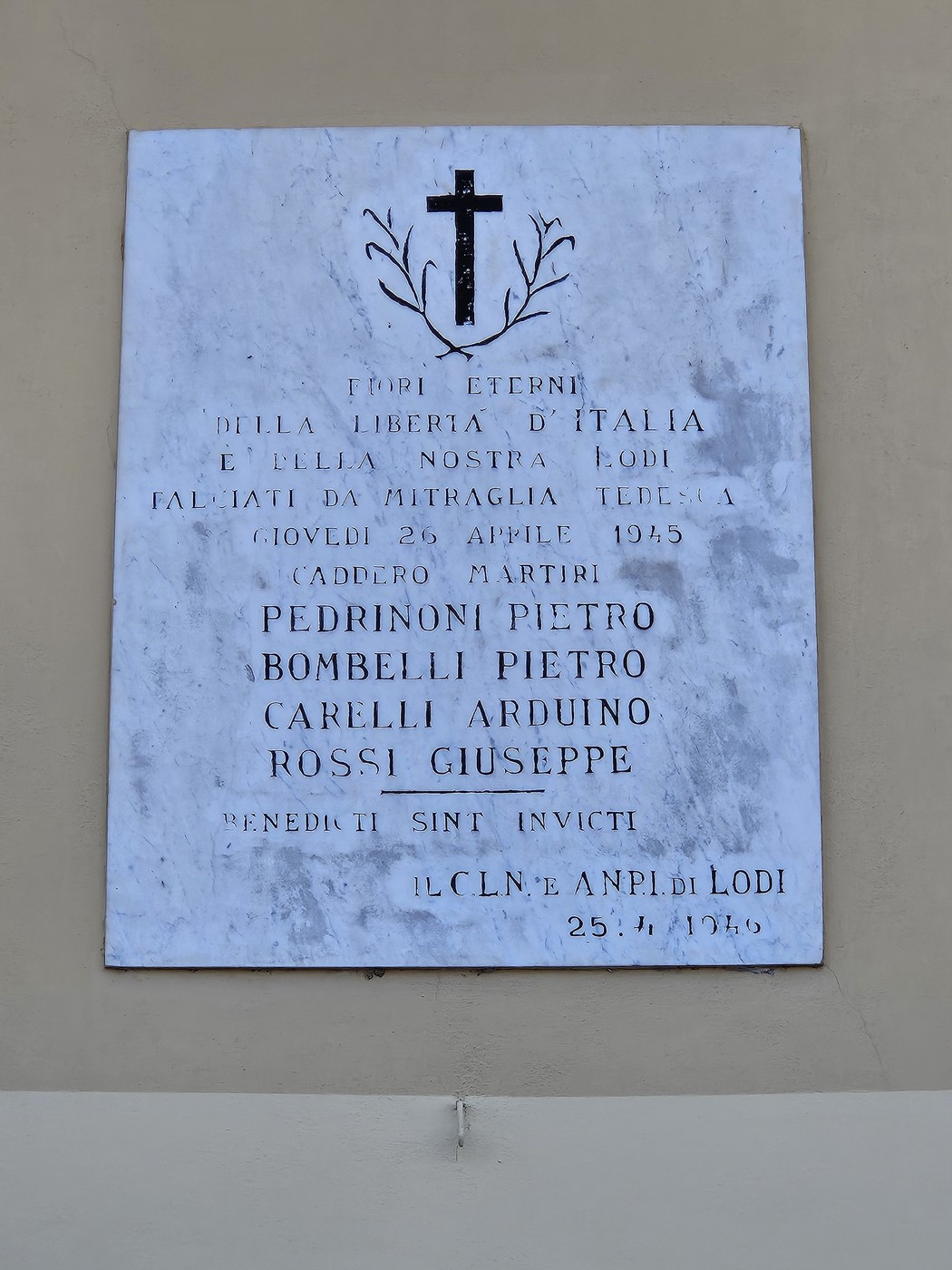 Lapide in marmo con croce scolpita e iscrizione, affissa su una parete esterna.
