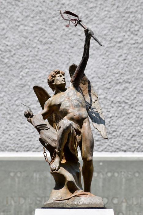 Statua bronzea di figura alata maschile con spada levata, su basamento con iscrizione, davanti a parete grigia.