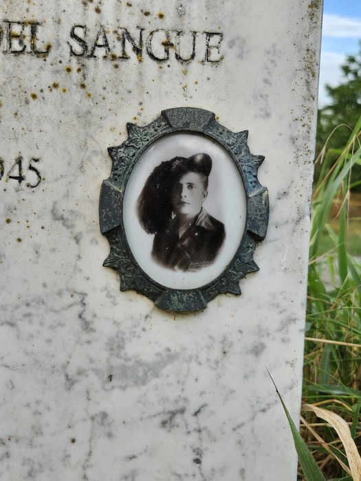 Cippo funerario con fotografia di un uomo in uniforme, circondato da vegetazione in un contesto rurale.