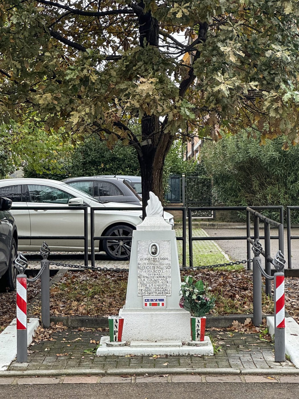 Cippo commemorativo dedicato a Bellotti, circondato da piante e veicoli, in un'area verde.