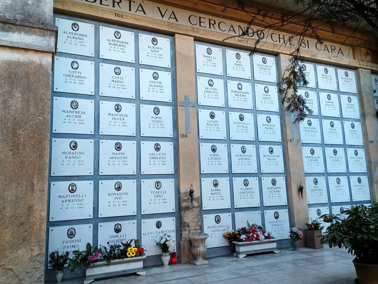 Arcate dei Caduti della Resistenza con lapidi bianche, fiori e una croce al centro, in un'atmosfera di commemorazione.