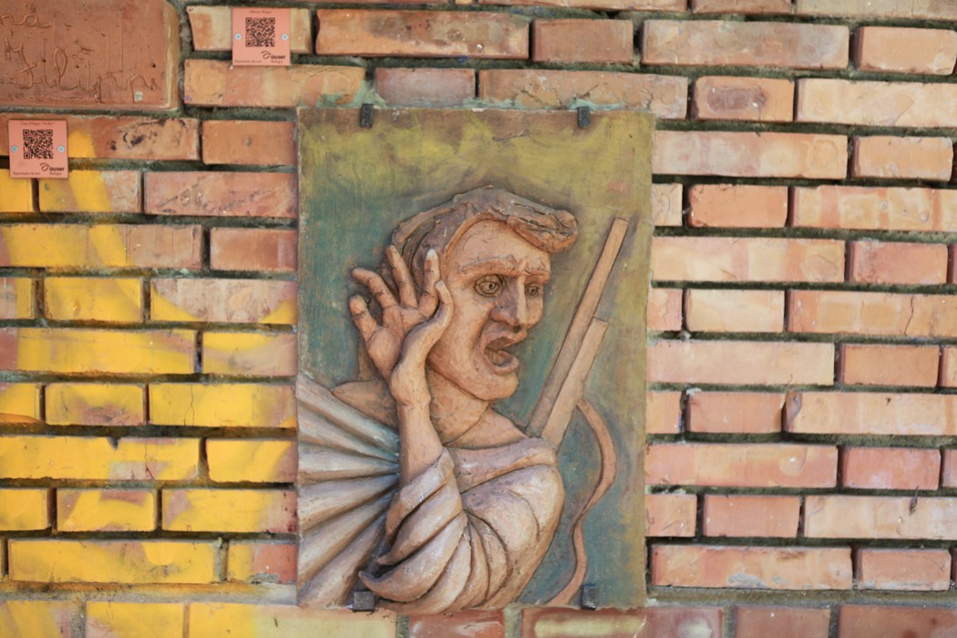 Scultura colorata rappresentante un volto umano in un muro di mattoni, simbolo di memoria storica.