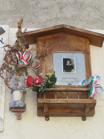 Targa commemorativa con foto e decorazioni floreali su un muro, in memoria di Maria Elina Borgis.