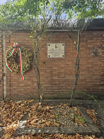 Lapide commemorativa decorata con una corona di foglie secche e nastri tricolore, circondata da foglie cadute.