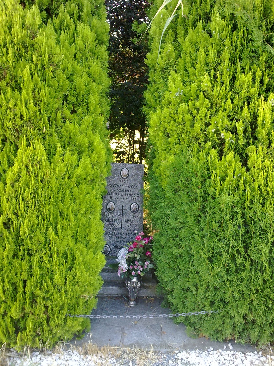 Cippo commemorativo dedicato ai fratelli Vecchi, circondato da siepi verdi e fiori.