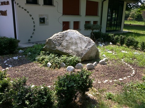 Monumento alle scuole Varini, situato in un'area verde, circondato da piante e sassi decorativi.
