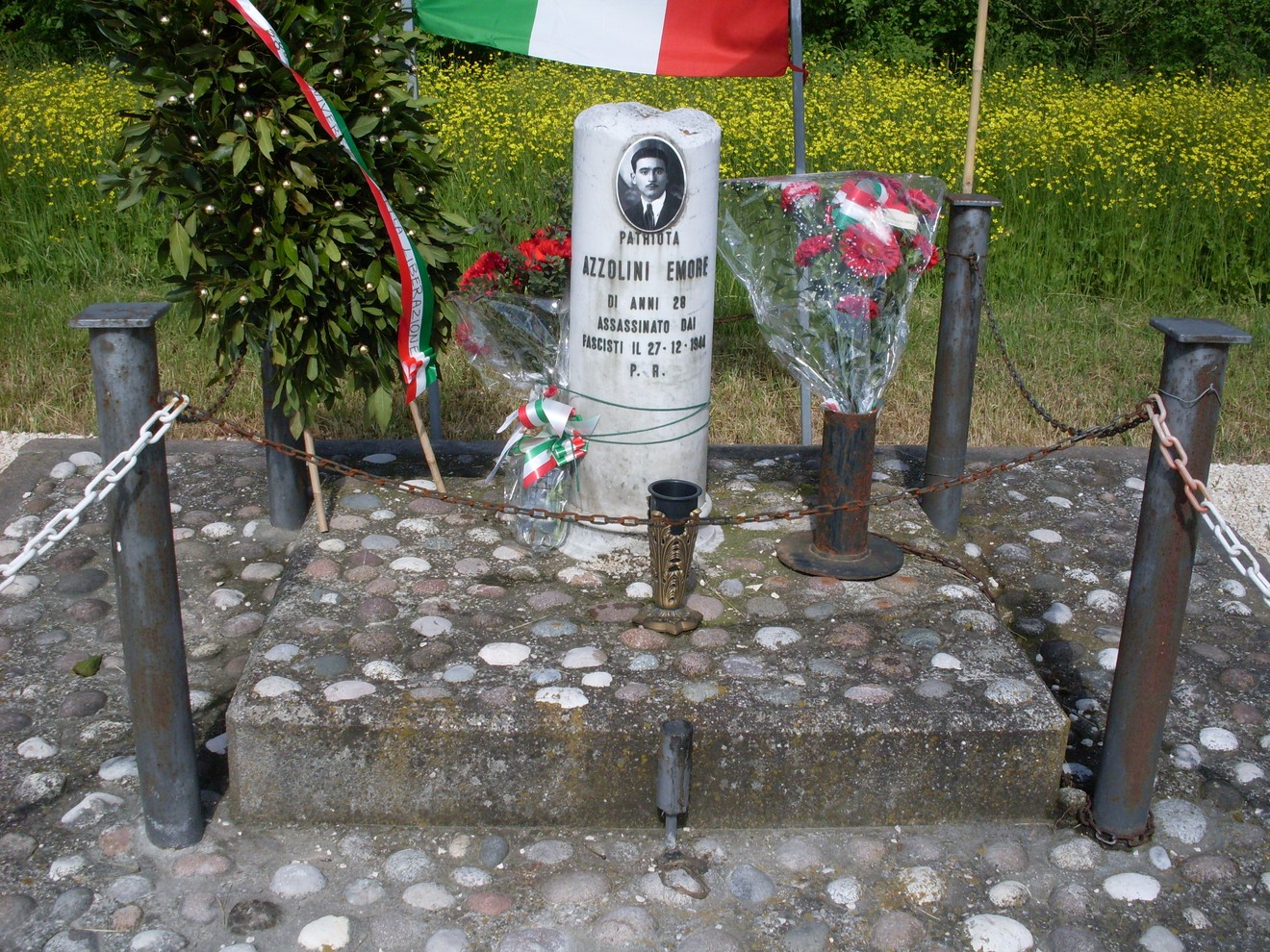 Cippo commemorativo di Azzolini, decorato con fiori e bandiere, in un contesto naturale di verde e fiori.