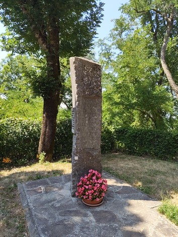 Cippo in pietra con iscrizione, su basamento in un giardino alberato, con vaso di fiori ai piedi.