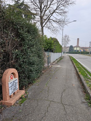 Cippo a Zani lungo un percorso pedonale, circondato da siepi e alberi, con una strada visibile in lontananza.
