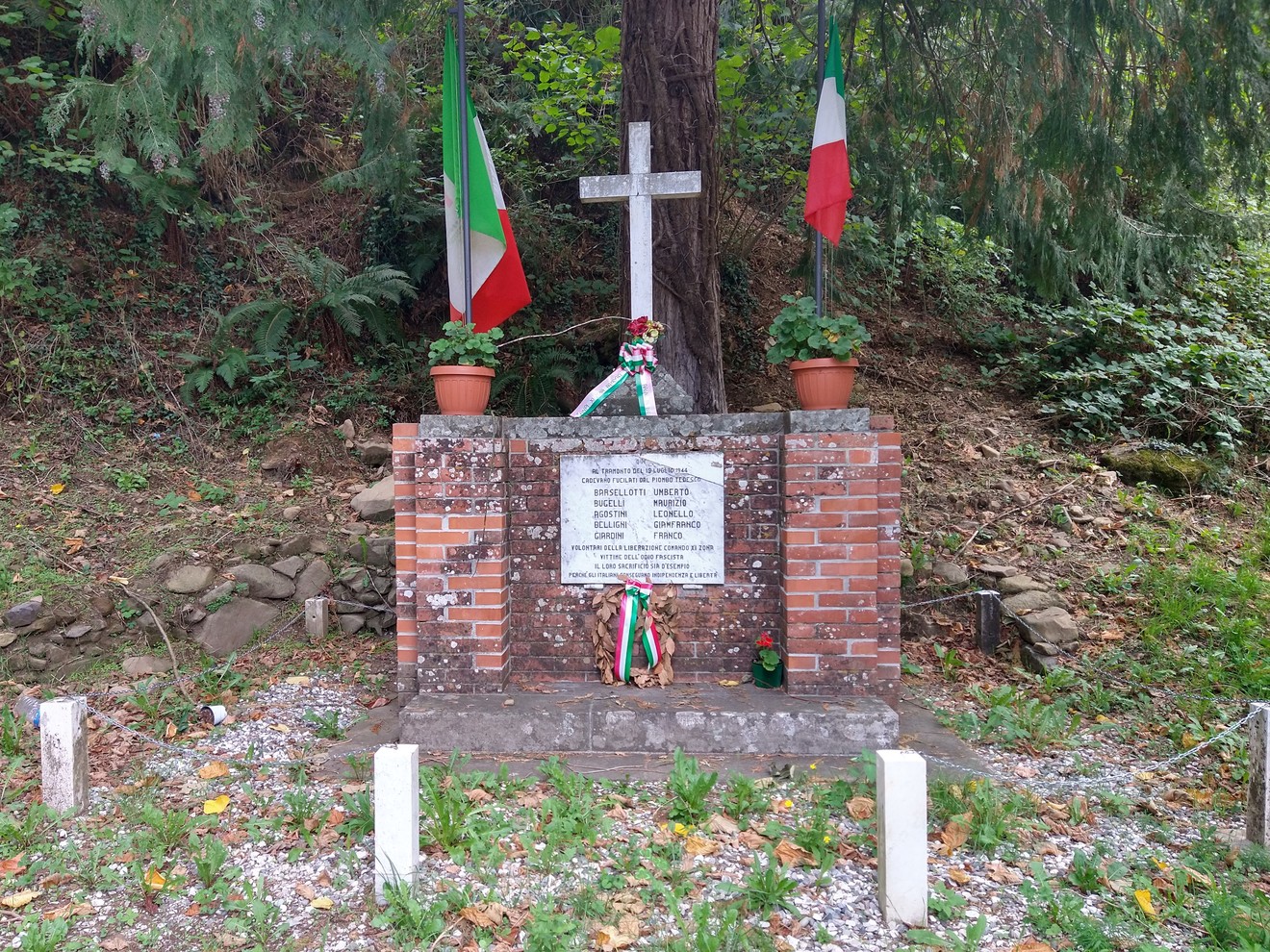 Monumento commemorativo a Barsellotti, Bugelli e altri, circondato da piante e bandiere italiane.