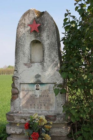 Cippo commemorativo di Saltini, con stella rossa e fiori, in un contesto rurale e verdeggiante.