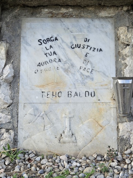 Cippo commemorativo con iscrizioni su una parete di pietra, circondato da ciottoli e vegetazione.
