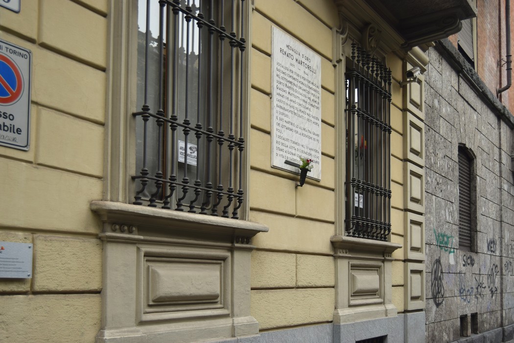 Lapide commemorativa affissa su una parete di un edificio, con finestre in ferro battuto e muratura storica.