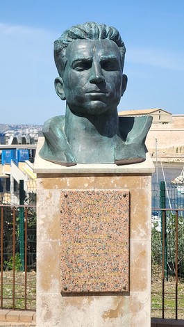 Il busto di un uomo su un piedistallo, immerso in un verde giardino con sfondo marittimo e cielo blu.