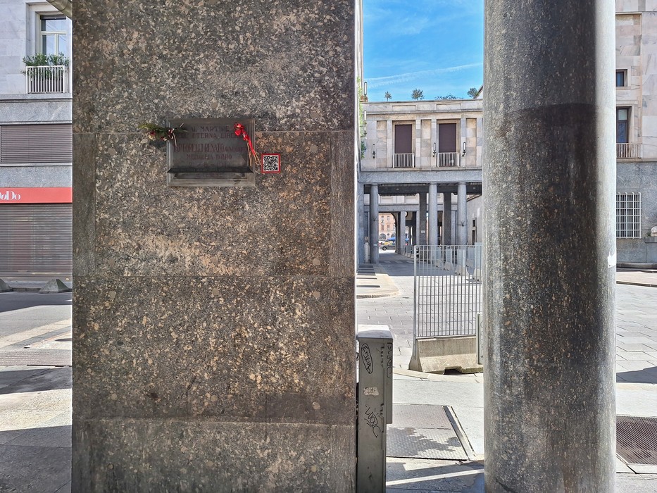 Lapide commemorativa su una colonna, affiancata da elementi architettonici e spazi urbani di Martorelli.