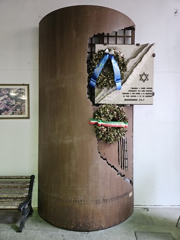Monumento alla Shoah con una struttura metallica e corone di fiori commemorativi appese.