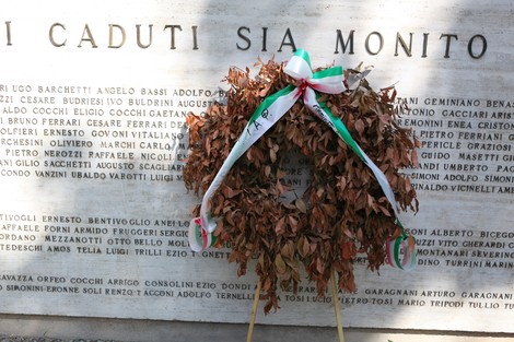 Monumento ai Caduti, omaggio floreale con foglie secche e nastri tricolore, collocato davanti a una stele commemorativa.