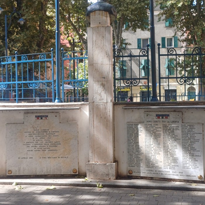 Due lapidi con iscrizioni ai lati di una colonna in pietra, su una piazza alberata con cancellata.