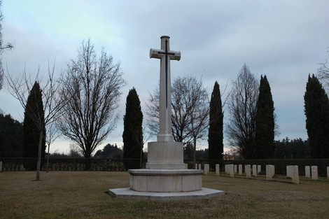 Cimitero di guerra del Commonwealth con una croce monumentale circondata da alberi e lapidi. Atmosfera solenne e tranquilla.
