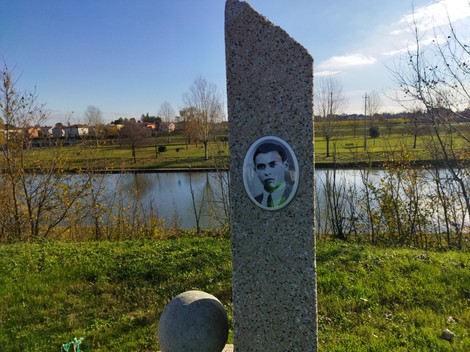Cippo a Fiocchi con foto commemorativa, immerso nella natura e vicino a un corso d'acqua.