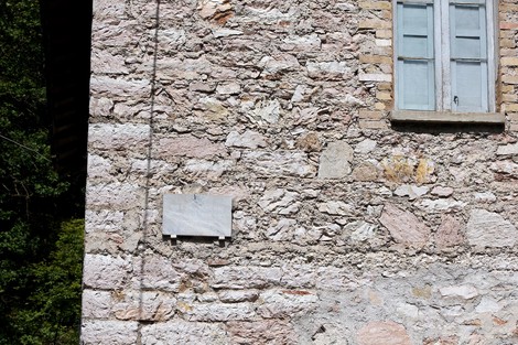 Lapide commemorativa su un muro di pietra, con finestre in legno sullo sfondo.