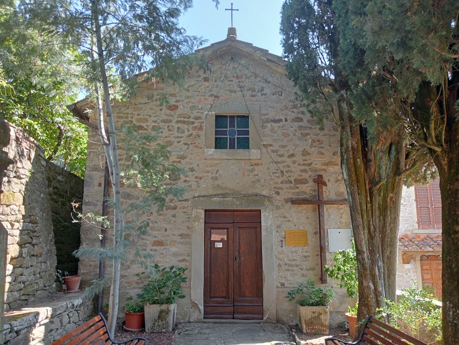 Chiesa in pietra con porte in legno, circondata da alberi e piante, in un contesto rustico e sereno.