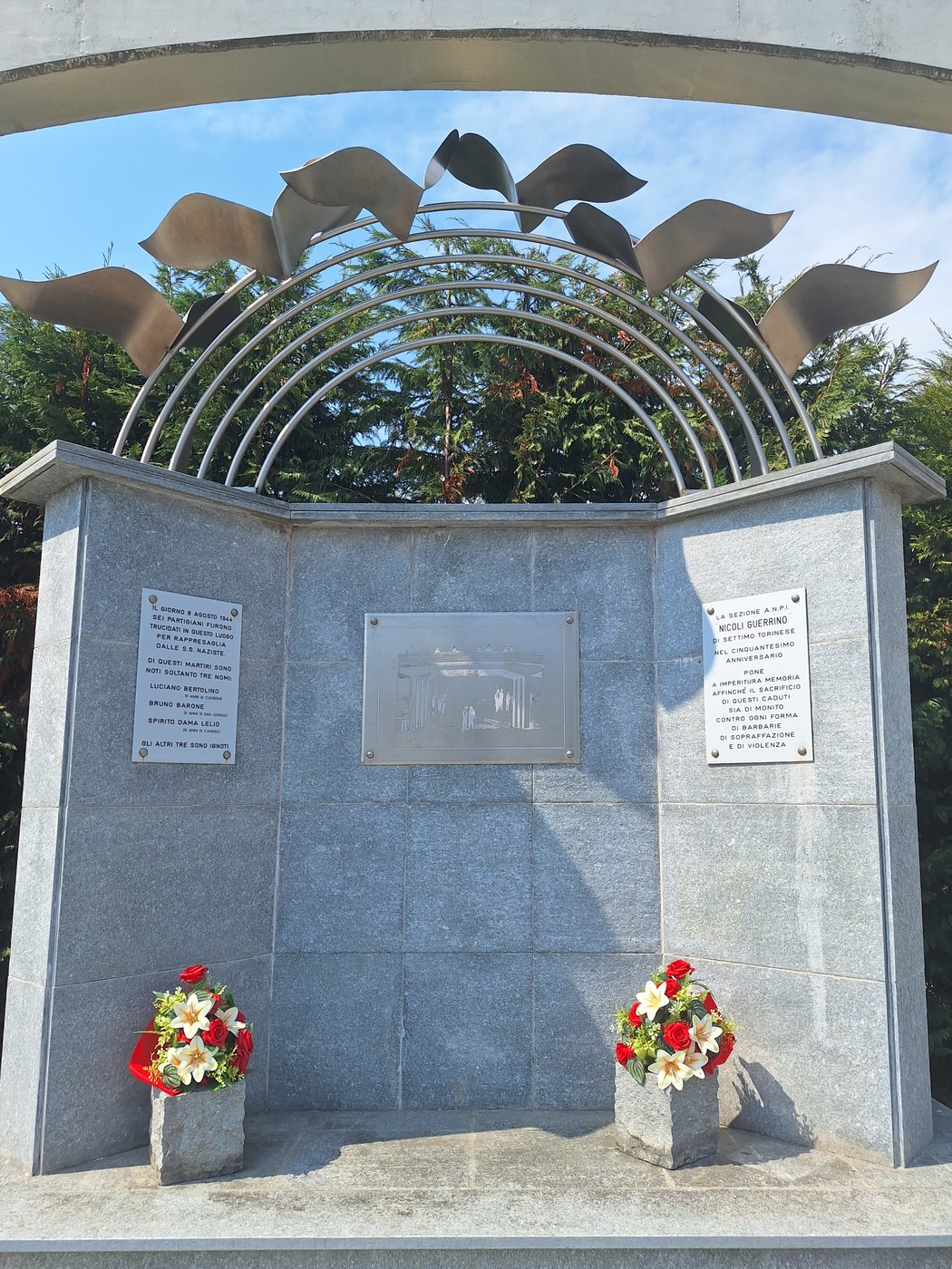 Monumento in granito con iscrizioni e arco di metallo decorato, addossato a una parete e ornato da fiori.