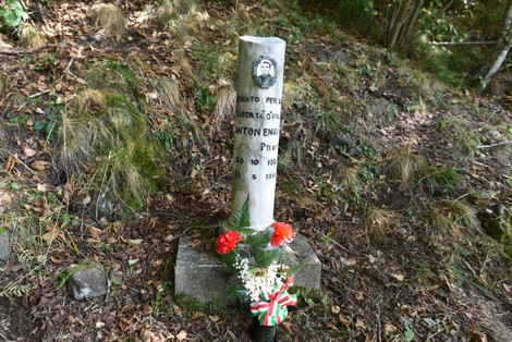 Cippo commemorativo in memoria di Anton Enzo, decorato con fiori, immerso nella natura.